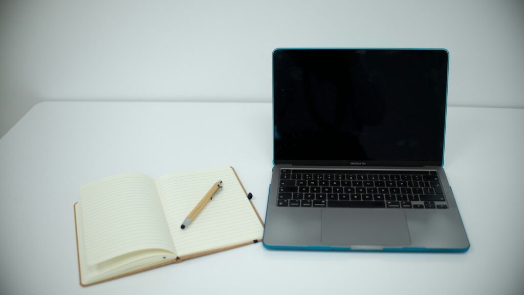 An open notebook and pen beside a laptop on a desk.
